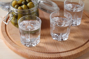 Glasses with vodka and a jar of cucumbers on a wooden plate.