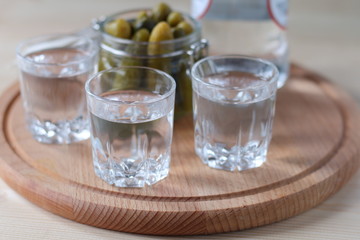 Glasses with vodka and a jar of cucumbers on a wooden plate.