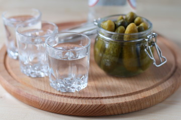 Glasses with vodka and a jar of cucumbers on a wooden plate.