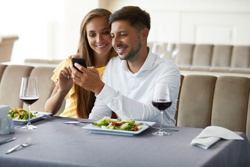 Couple In Love Looking On Phone Having Dinner In Restaurant.