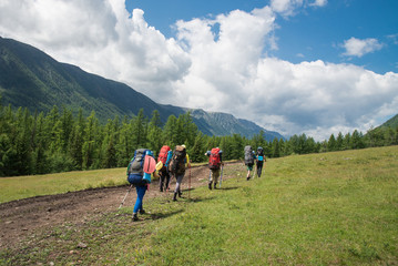 Group of travelers with backpacks walk along a trail towards a mountain ridge by sunny day. Backpackers and hikers style, active leisure