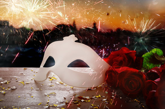 Image of elegant venetian mask and red roses over wooden table in front of blurry Venice background
