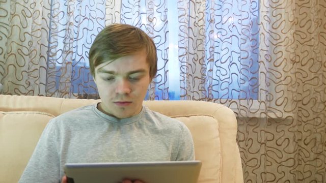 Handsome man is using a tablet while lying on couch at home. Young man working with laptop at home