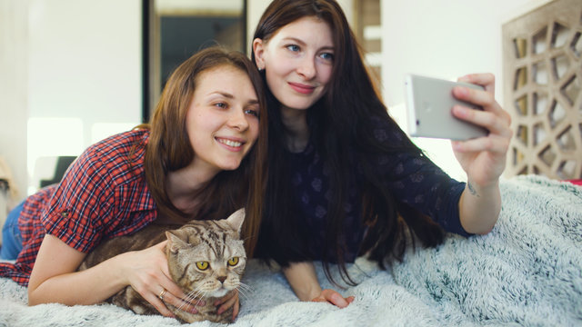 Two happy women friends lying in bed and making selfie with cat and have fun on bed at home