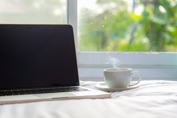 The laptop working on the white bed and coffee in the window on rainy day, Fresh morning holiday.  Lifestyle Concept. soft and select focus.