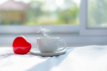 Fresh Cozy morning coffee on the white bed with red heart shape near window in the sunny day, green nature background.  Lifestyle & Valentine Concept