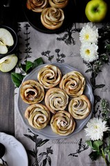 On a wooden table on a platter, apples are baked in the form of roses. The table is decorated with cotton napkins, fresh apples and flowers