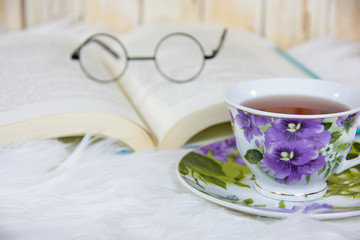 hot tea in vintage pansy floral pattern tea cup on saucer and white fur with old-fashioned glasses on open book