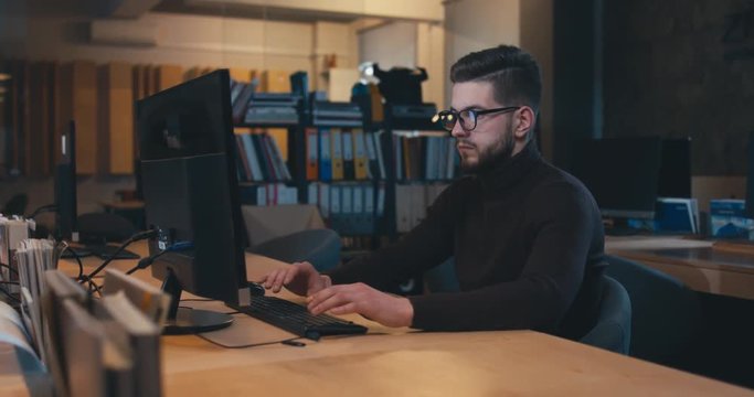 Young adult professional Caucasian male employee working late at the office, typing something on a computer. 4K UHD