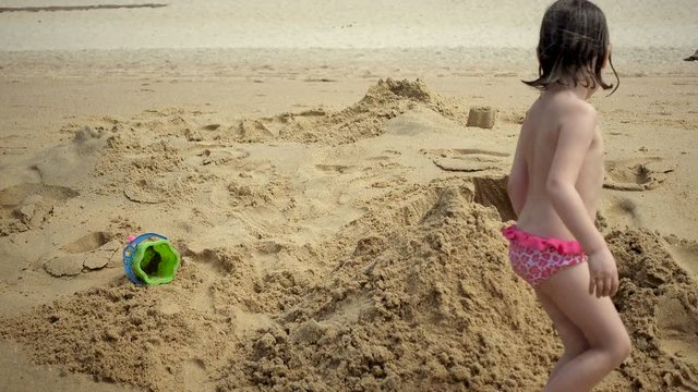 A white-haired girl of European appearance, four or five years old, plays on the beach. The child builds a sand castle.
