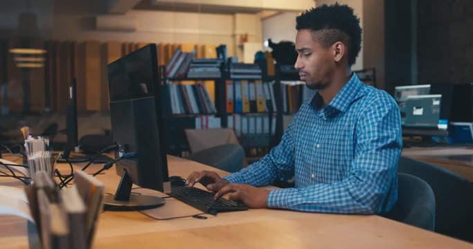 Young adult professional African American black male employee working late at the office, typing something on a computer. 4K UHD