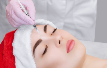 Obraz na płótnie Canvas The cosmetologist makes the Botulinum toxin injection on the face skin of a beautiful, young woman in the Santa Claus hat. New Year's and Cosmetology concept