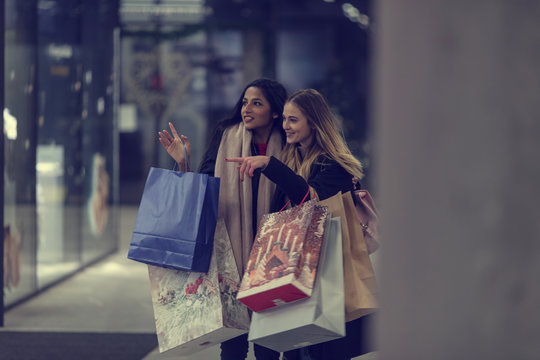 Two cute young adolescent women, heavy loaded with christmas gifts,  shopping, while walking along store windows and choosing and admiring the items. 