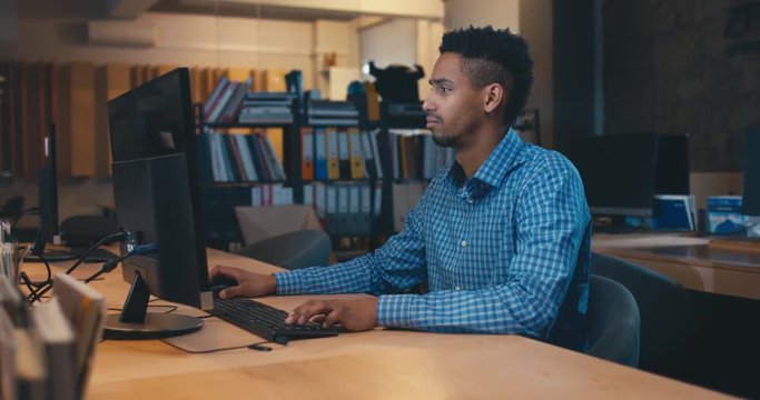 Young adult professional African American black male employee working late at the office, typing something on a computer. 4K UHD