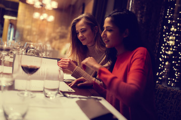 Two adolescent women going through the menue  in a fancy restaurant, while drinking a glass of red wine and laughing and talking, maybe gossiping