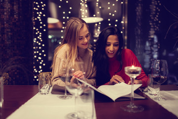 Two adolescent women going through the menue  in a fancy restaurant, while drinking a glass of red wine and laughing and talking, maybe gossiping