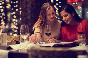 Fototapeta premium Two adolescent women going through the menue in a fancy restaurant, while drinking a glass of red wine and laughing and talking, maybe gossiping