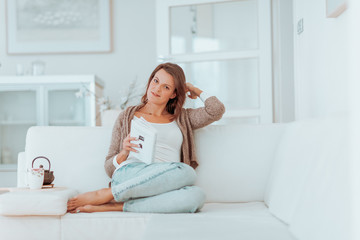 young woman in her late twenties sitting and laying on a lether white sofa in a cosy interier of her bright home and drinking a cup of tea and reading a book, totaly relaxed