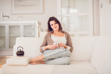 young woman in her late twenties sitting and laying on a lether white sofa in a cosy interier of her bright home and drinking a cup of tea and reading a book, totaly relaxed