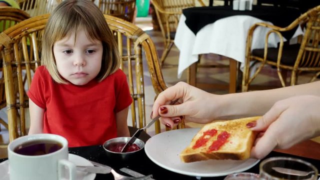A child, a Russian girl, 4-5 years old, refuses to eat a toast with jam for breakfast. She sits at the table.