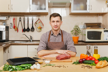 Handsome caucasian young man in apron sitting at table with vegetables, cooking at home preparing meat stake from pork, beef or lamb, in light kitchen with wooden surface, full of fancy kitchenware.