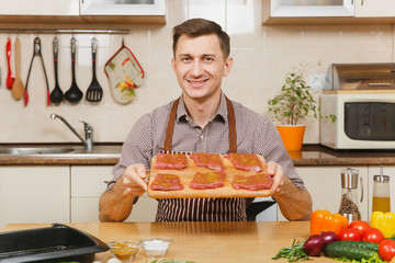 Young man in apron sitting at table with pieces of meat on wooden cutting board, vegetables,...