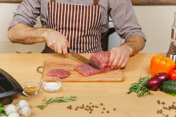 Close up caucasian young man in apron sitting at table with vegetables, cooking at home preparing meat stake from pork, beef or lamb, in light kitchen with wooden surface, full of fancy kitchenware.