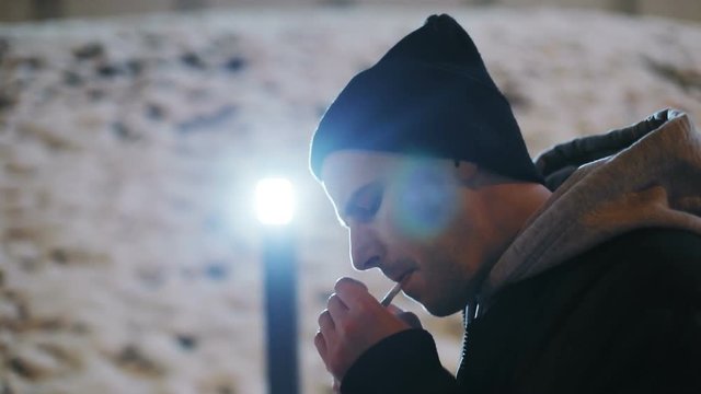 caucasian man lighting cigarette outside street winter calm male person evening warm clothes black cap smoking lighter tobacco smokes weed addiction vapor drugs healthcare alone walking city side view