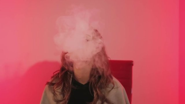 cute girl hovering vaporizer on the chair with red backrest, which is located in room with red lighting on the background