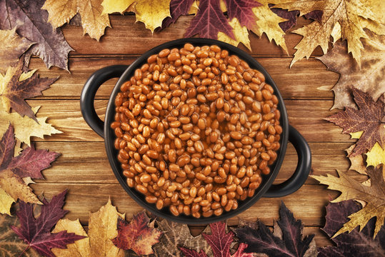 Overhead photo of a pot of baked beans on a wooden table and autumn fall leaves in the background.