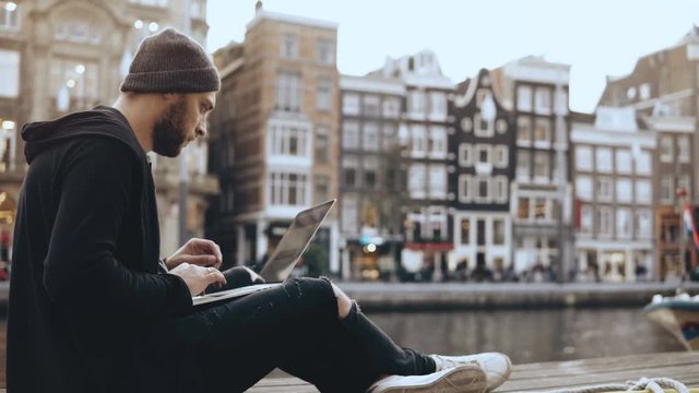 4K Young creative businessman uses laptop outside. Successful man working near beautiful river embankment, old town.