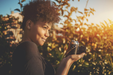 Appealing serious black curly female with vintage film camera is taking pictures in sunny garden; charming young African American female with retro photo camera in city parkland photographing plants