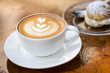 Cup of coffee with flower latte art on wooden table. Chu cream puff recipes in background. Warm morning soft light effect added. - obrazy, fototapety, plakaty