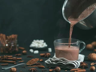 Wall murals Chocolate Pouring tasty hot chocolate cocoa drink into glass mug with ingredients on black table. Copy space Dark background. Low key.