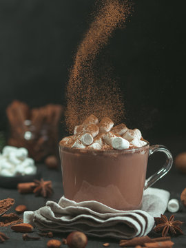 Hand sprinkled cinnamon powder on glass mug with hot chocolate cocoa drink. Copy space. Dark background. Low key. Winter food and drink concept.