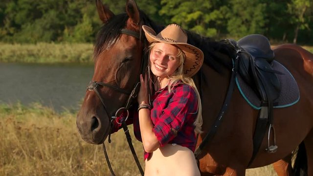 Portrait of a young girl in a cowboy hat near a horse, close-up. Slow motion. Young woman near a horse in evening sunset light.