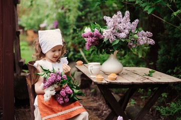 kid girl at garden tea party in spring day with bouquet of lilacs (syringa), rustic wooden table and vintage dress