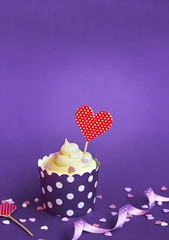 Vanilla cupcake with small decorative hearts and red heart topper, in violet baking paper cup, against violet background Cupcake for Valentine's day with copy space