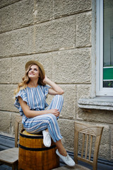 Portrait of a good-looking young woman in striped overall posing with her hat and sitting on a wooden barrel outdoor.