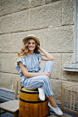 Portrait of a good-looking young woman in striped overall posing with her hat and sitting on a wooden barrel outdoor.