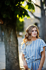 Portrait of a very attractive young woman in striped overall posing with her hat on a pavement in a town with trees in a background.