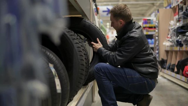 Young man in supermarket choosing automobile tyres standing on the shelves