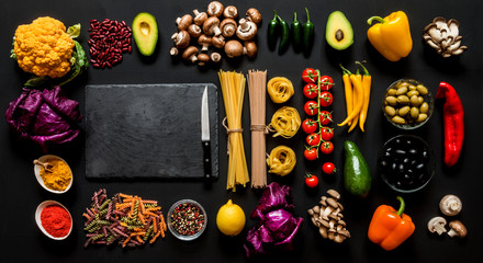 Different colorful Fresh raw ingredients for healthy vegetarian cooking on a black background with blackboard and free copy space. Flat lay, top view.