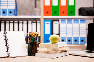 Green apple on pile of books next to a notebook and pencils on table with a blurred white board in the back. School concept