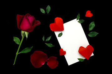 Top view (flat lay) image of white paper note, red rose, green leaf, red origami paper heart on black baclground. Valentine's day concept 