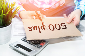 Businessman is cutting cardboard with scissors. Documents, calculator, telephone on the table. It cuts one half. Cuts in the Polish social program 500 plus, problems with 500+. The man cuts the plus.