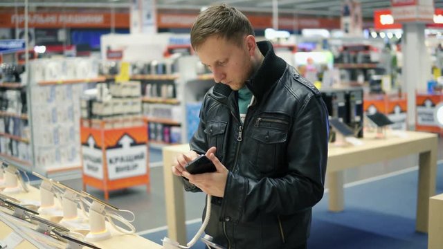 Young man in black leather jacket is choosing a new mobile phone in a shop, checking how it works