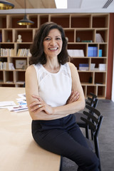 Middle aged female creative sitting on desk smiles to camera