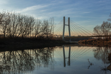 Lanscape with Siekierkowski suspension bridge across the Vistula River and its reflection in water among bare trees in colorful light of the setting sun