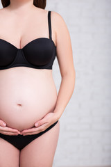 close up of pregnant woman in black underwear touching her belly over white wall